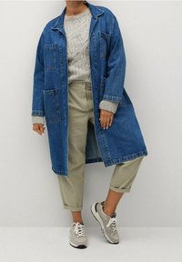 Person som bär en lång blå denimjacka över en grå stickad tröja, beige uppvikta byxor och grå sneakers, stående mot en enkel bakgrund.