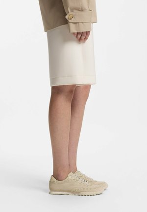 Person iført beige sneakers, knælang cremefarvet nederdel og beige trenchcoat, stående på siden mod en hvid baggrund.