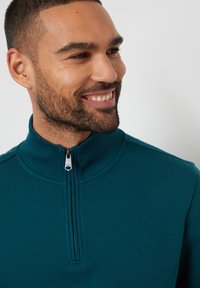 Tyrkysová fleecová mikina s vysokým límcem, polovičním zipem vpředu a jemnou vyšitou dekorací na hrudi, která má měkkou texturu a ležérní design.