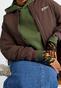 Osoba v hnědé bundě New Balance přes zelenou mikinu, s dlouhými rukávy s tygřím potiskem, modrou džínovou sukní a béžovými teniskami, sedící s rukama odpočívajícími.