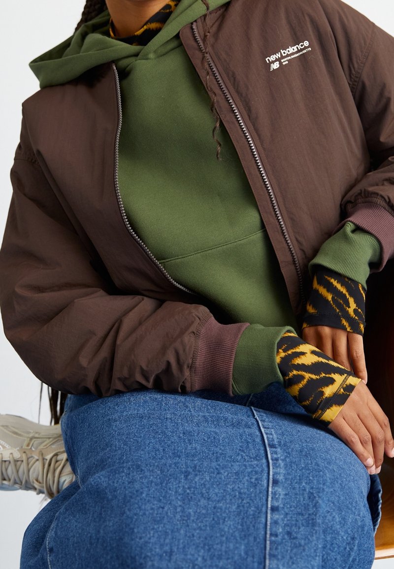 Osoba v hnědé bundě New Balance přes zelenou mikinu, s dlouhými rukávy s tygřím potiskem, modrou džínovou sukní a béžovými teniskami, sedící s rukama odpočívajícími.
