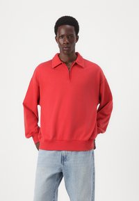 Pullover rosso con design a collo con zip, vestibilità rilassata e polsini a costine. Abbinato a jeans azzurri chiari, con una texture denim classica.