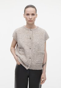 Beige korte mouwen cardigan van fluffy materiaal met zilverkleurige knopen en twee voorzakken. Glad textuur, relaxte vorm en subtiele details.