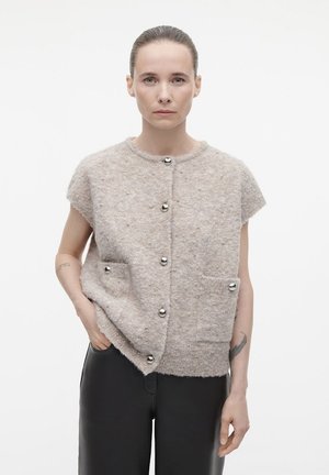 Beige korte mouwen cardigan van fluffy materiaal met zilverkleurige knopen en twee voorzakken. Glad textuur, relaxte vorm en subtiele details.