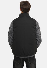 Veste sans manches noire matelassée avec un col montant, des manches grises texturées et un ourlet élastique. La vue de dos met en avant un matériau lisse et un contraste de couleurs distinct.