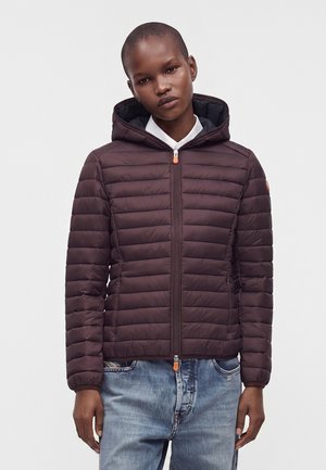 Person iført en mørkebrun polstret hættetrøje med lynlås foran og lyseblå jeans, stående mod en ensfarvet lys baggrund.