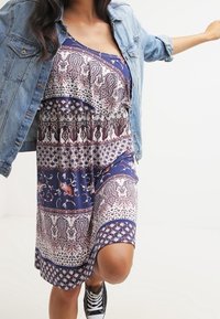 Vestido azul con estampado y volantes en capas, con diseños de paisley y flores. Combinado con una chaqueta de denim clara y zapatillas negras.