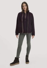 Paarse fleece-jack met een ritssluiting en bruine accenten, gecombineerd met olijfgroene leggings en zwarte enkellaarsjes met een ribbelzool.