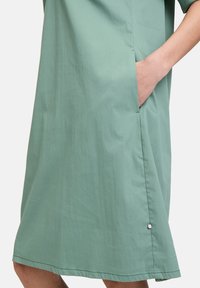 Mintgrünes Kleid mit lockerer Passform, ausgestattet mit einer Seitentasche und einem kleinen silbernen Akzent am Saum. Glatte Textur, knielange Schnittform.