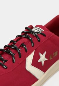 Red suede sneaker with black laces, white star logo, and beige sole. Features "ALL STAR" text in gold on the side. Smooth texture.