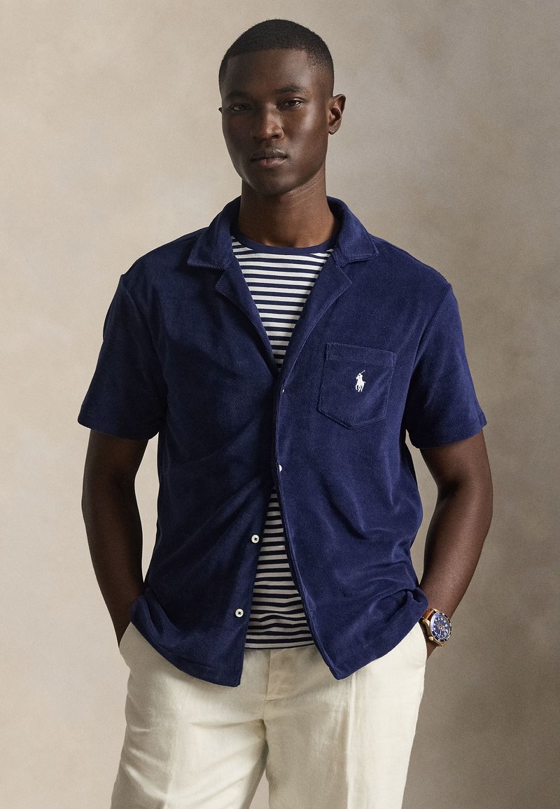 Σκούρο μπλε κοντομάνικο πουκάμισο από απαλό ύφασμα με τσέπη, διακοσμημένο με λευκό λογότυπο. Φοριέται πάνω από ριγέ μπλούζα, συνδυασμένο με ανοιχτόχρωμο παντελόνι.