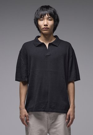 Černé žebrované polokošile s volnějším střihem, se žlábkovým límečkem a mírně rozepnutým předním dílem. Látka je texturovaná a měkká.