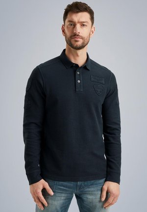Navy long-sleeve polo shirt with a collar, textured fabric, three-button placket, and embroidered patches on sleeves. Worn with light blue jeans.