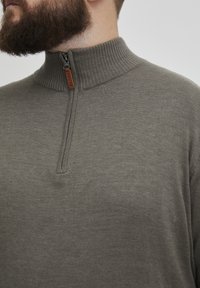 Jersey de cuello alto gris con cremallera en textura acanalada, que presenta un cuello alto y una lengüeta de cuero marrón en contraste con texto en relieve.