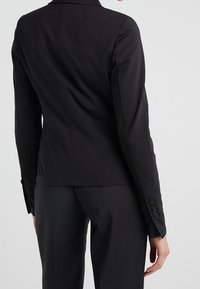 Donna che indossa un blazer nero aderente con quattro bottoni sul polsino, abbinato a pantaloni neri coordinati, mostrata di schiena.