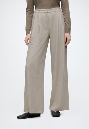 Person trägt beige weite Hosen, braues Langarmshirt und schwarze flache Schuhe, steht vor einem schlichten hellen Hintergrund.