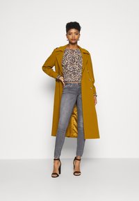Casaco longo mostarda com botões duplos sobre uma blusa floral, combinado com jeans skinny cinzentos e sandálias de salto alto pretas.