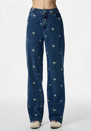 Jeansy z wysokim stanem w ciemnym odcieniu niebieskiego z drobnym, białym kwiatowym haftem, noszone z beżowymi sneakersami, pokazane od talii do stóp.