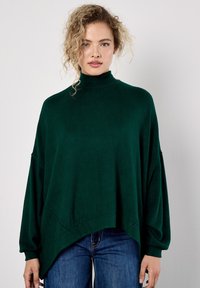 Frau mit lockigem blonden Haar, die einen dunkelgrünen, übergroßen Rollkragenpullover und bluejeans vor einem einfachen hellen Hintergrund trägt.
