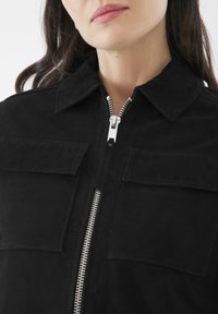 Giacca nera in suede con colletto, zip argentata e due tasche frontali. Il tessuto ha una texture liscia e opaca ed un design aderente.