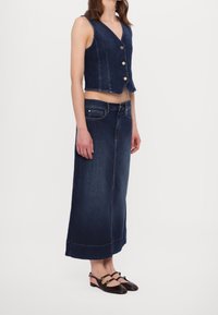 Džínová vesta s krátkým designem, pěti knoflíky, spárovaná s dlouhou tmavou džínovou sukní. Obuv zahrnuje černé sandály s pásky s detaily.