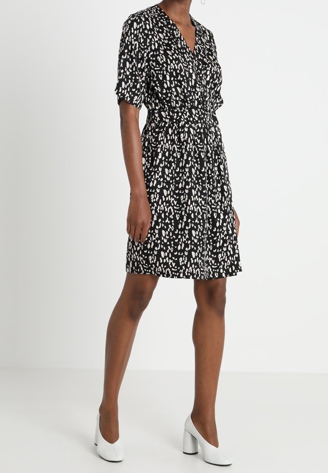 Vestido negro con patrón salpicado blanco, escote en V, mangas cortas, cintura ceñida y dobladillo acampanado. Combinado con zapatos de tacón cuadrado blancos.