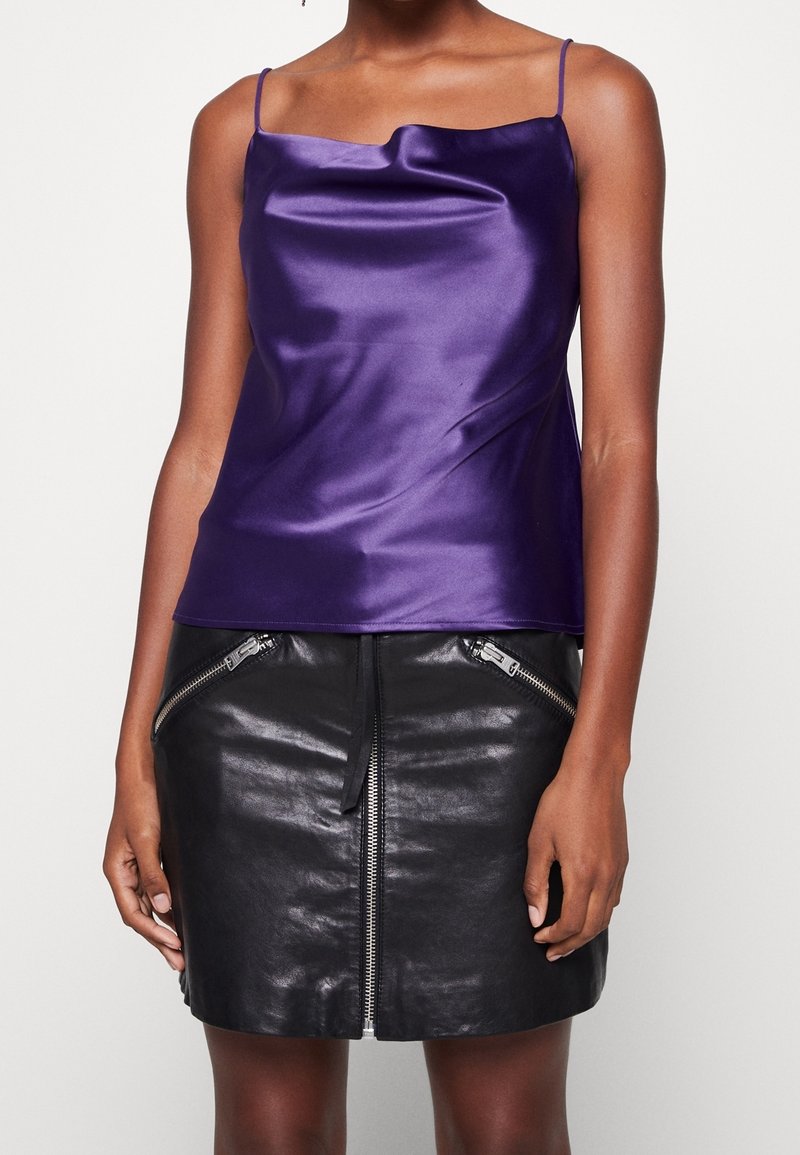 Femme portant un caraco en satin violet brillant avec de fines bretelles et une mini-jupe en cuir noire avec des détails de fermeture éclair à l'avant.