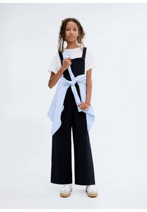 Salopette noir à jambes larges superposée sur un t-shirt blanc, avec une chemise rayée bleu clair nouée à la taille. Baskets blanches avec semelles marron clair.