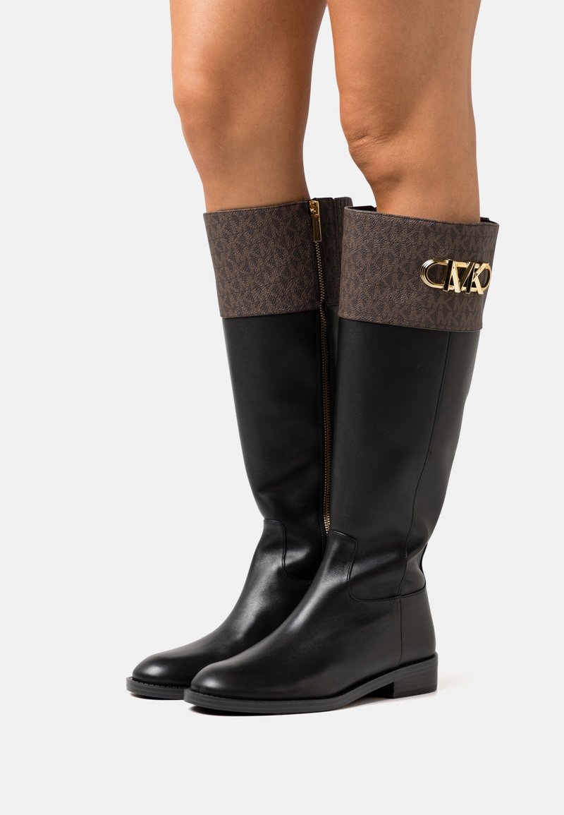 Bottes en cuir noir montantes jusqu'au genou avec des revers marron à motifs, fermeture éclair dorée sur le côté intérieur, et détail du logo "MK" doré sur le revers extérieur.