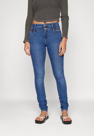 Slim-fit blue jeans with double-button waist and zipper pockets, worn with brown cropped long-sleeve top and white-strap platform sandals.