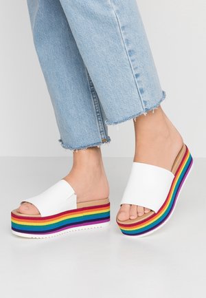 Pies con sandalias tipo chancleta blancas con suelas de plataforma gruesas con rayas de arcoíris y jeans cortos desgastados azul claro sobre un fondo blanco.