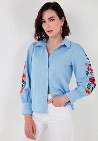 Light blue button-up shirt with floral embroidered patterns on the sleeves. Features structured collar and gathered cuffs. Smooth fabric texture.
