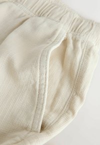 Zbliżenie na kremowe spodnie z fakturowanej tkaniny z elastycznym pasem i widocznym przeszyciem kieszeni bocznej.
