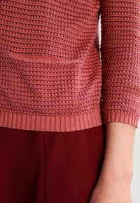 Rosa texturerad tröja med rund halsringning och ribbade manschetter, med en framficka; kombinerad med mörkröda byxor.