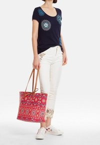 Camiseta marina con patrones decorativos azules, pantalones blancos con bordados florales, bolsa de mano roja con estampado y zapatillas blancas.
