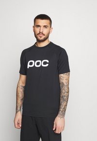 Černé sportovní tričko krátkého rukávu s hladkým povrchem a velkým bílým logem "poc" na přední straně. Obsahuje kulatý výstřih a raglánové rukávy.
