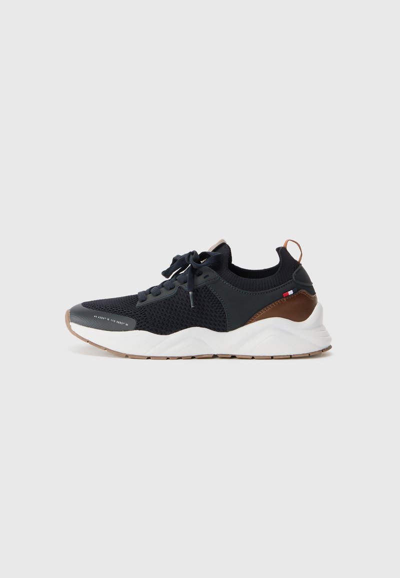 Black athletic sneakers with a textured mesh upper, curved white sole, and brown accents on the heel and sides. Features lace-up design.