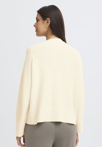 Grietos spalvos megztinis su laisvu siluetu, nuleistais pečiais ir nertais kraštais. Yra minimalus dekoras ir lygus audinys nugarinėje pusėje.