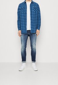 Man i blå rutiga skjortor över vit t-shirt, urtvättade blå jeans uppvikta vid fållarna, och vita sneakers mot en enfärgad bakgrund.