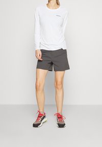 Valkoinen pitkähiha-shirti, jossa on pieni logo rinnassa, yhdistettynä harmaisiin shortsit ja harmaisiin lenkkarisi, joissa on punaiset nauhat ja vihreät yksityiskohdat.