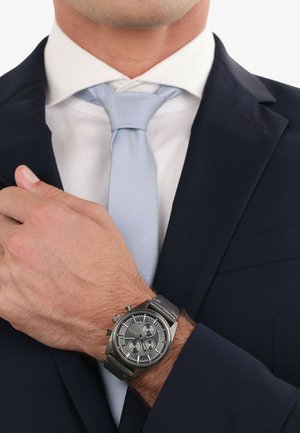 Silberner Chronographuhr an einem Handgelenk, mit einem grauen Lederarmband, runder Zifferblatt und drei Hilfszifferblättern, kombiniert mit einem dunklen Anzug und einer hellblauen Krawatte.