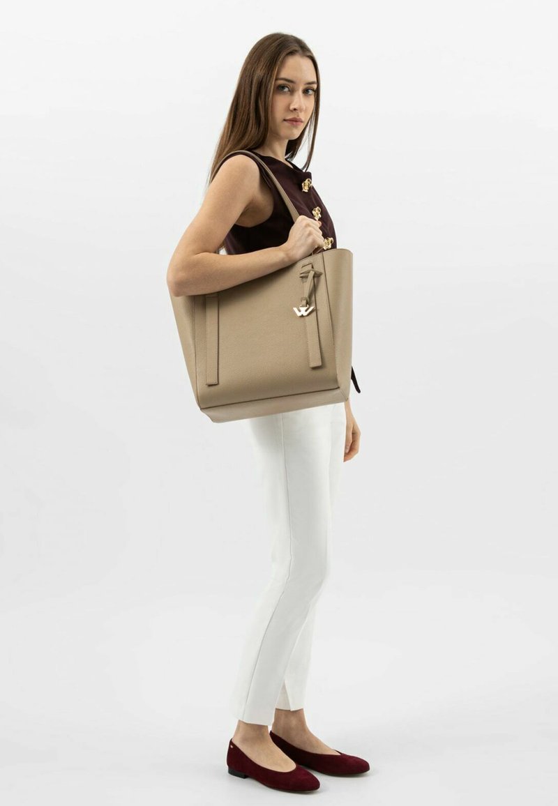 Beżowa skórzana torba podręczna o strukturowanym kształcie, wyposażona w krótki uchwyt i dekoracyjne logo. Modelka ma na sobie białe spodnie i czerwone baleriny.