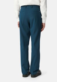 Heren gepaste broek van diep teal stof, met een gladde textuur, opvallende naden en twee achterzakken, gecombineerd met zwarte schoenen.