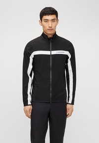 Veste zippée noire avec des rayures latérales blanches et un logo, présentant un col montant et une coupe ajustée, fabriquée en tissu lisse et léger.