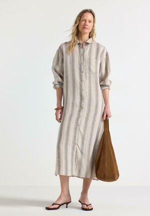 Kvinde iført en langærmet, knappet kjole med striber, der holder en brun ruskinds taske, stående i et minimalistisk hvidt rum.