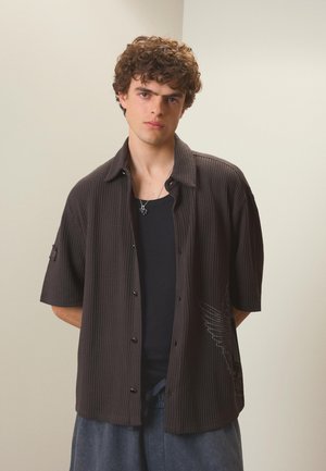 Jeune homme aux cheveux bouclés porte une chemise à manches courtes texturée de couleur sombre avec des boutons par-dessus un top noir, debout devant un fond clair uni.