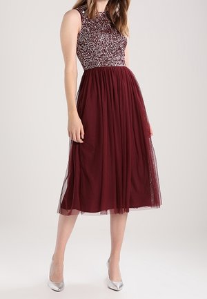 Mujer con vestido midi burdeos sin mangas con corpiño de lentejuelas y falda de tul, combinado con tacones altos plateados.