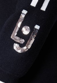 Il tessuto nero presenta un design di logo lucido in paillettes argento, con forme curve e punti, che aggiungono texture alla superficie.