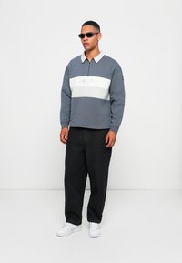 Ung mand iført mørke solbriller, en blå-grå zip-up sweatshirt med hvid stribe, sorte bukser og hvide sneakers, som står mod en hvid baggrund.