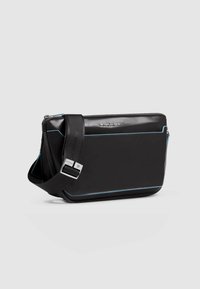 Bolso mensajero sintético negro con costuras decorativas en azul, cierre de cremallera y correa ajustable. Presenta un logo en la parte frontal. Forma rectangular.